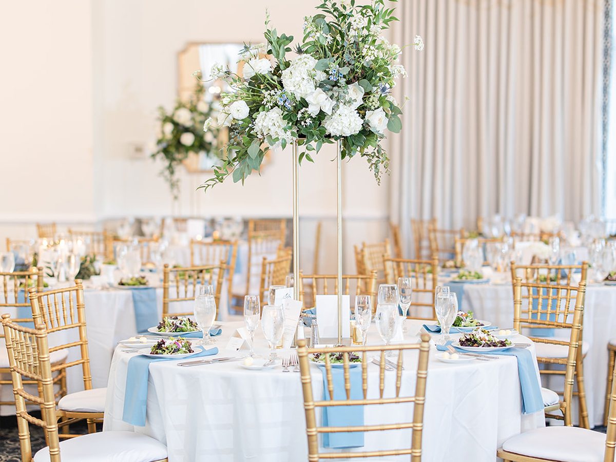 The Carolina Ballroom at Providence Country Club set for a wedding reception, with gold chairs, blue napkins and tall white floral centerpieces